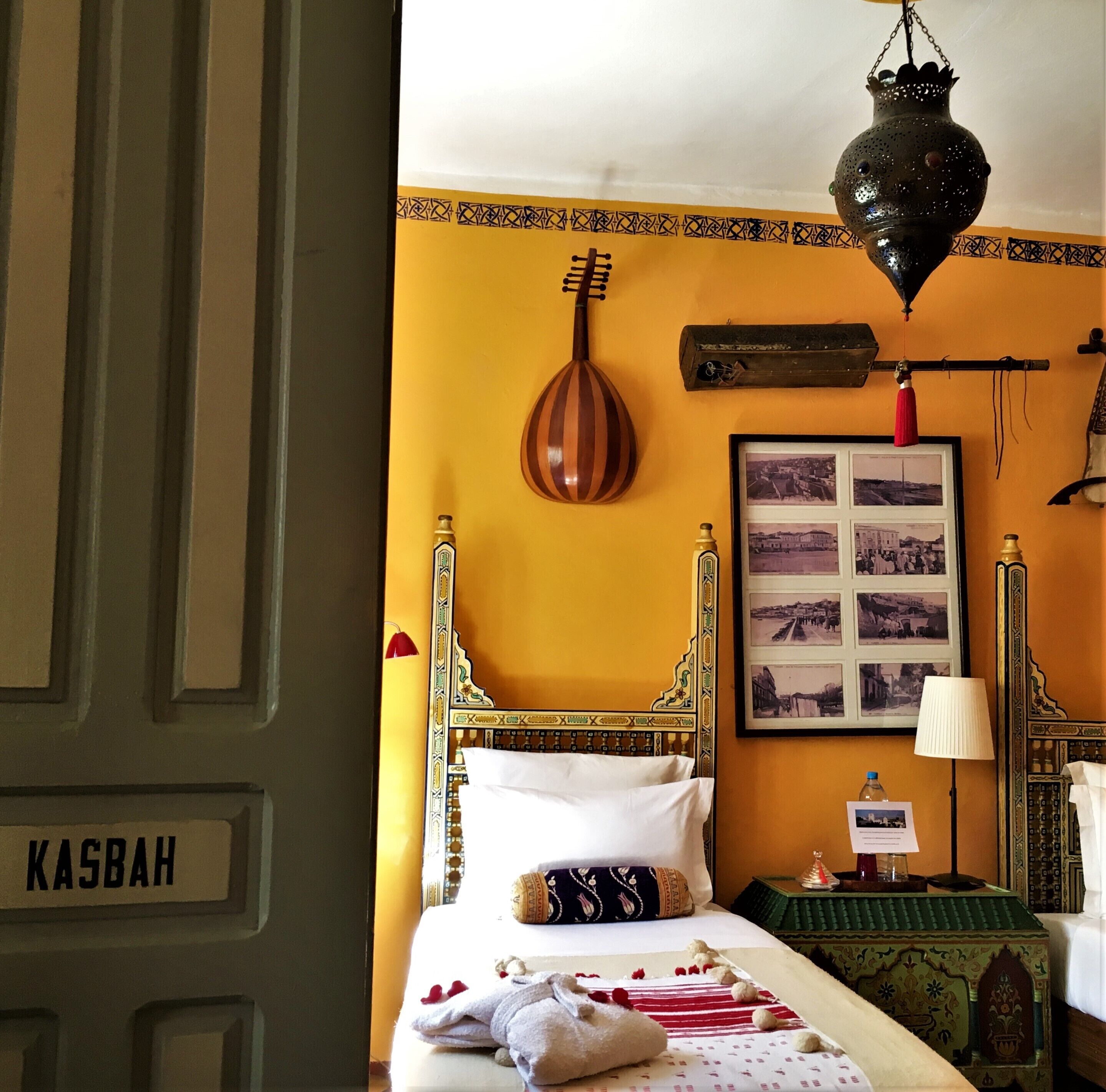 Una habitación con cama, lámpara colgante, un cuadro y una mesita con una botella.