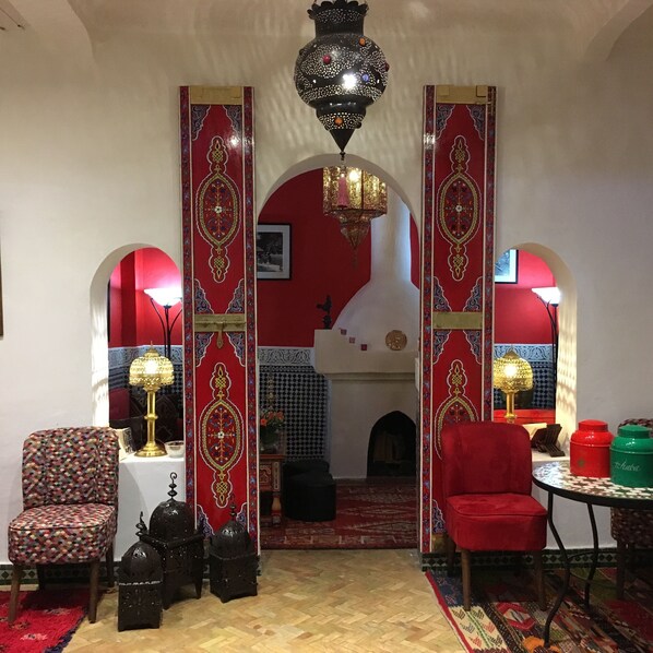 A room with a red and white patterned archway, a chandelier, a patterned armchair, a red chair, a small table with a lamp, and a patterned rug. - Dar Sultan (Tangier)