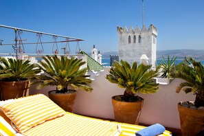 A balcony with a bed, potted plants, and a view of a castle and sea. - Dar Sultan (Tangier)