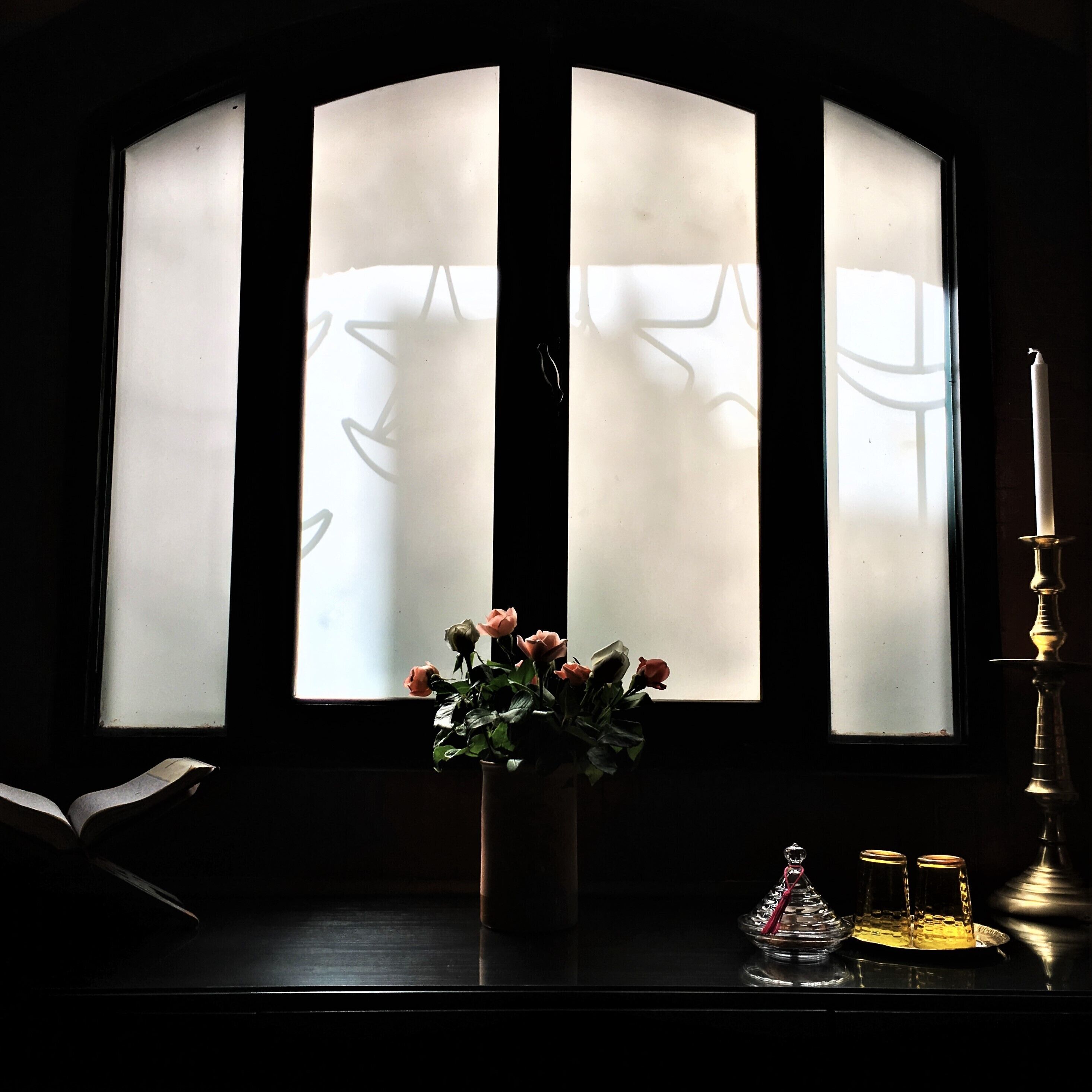 A dark room with a window divided into three sections, a vase with flowers, a candle, and a book on a table.