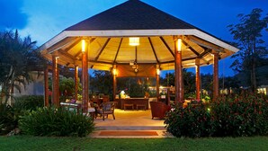 Un espace extérieur abrité, doté d’une table et de chaises au centre, et entouré d’une végétation luxuriante et d’un jardin bien entretenu.