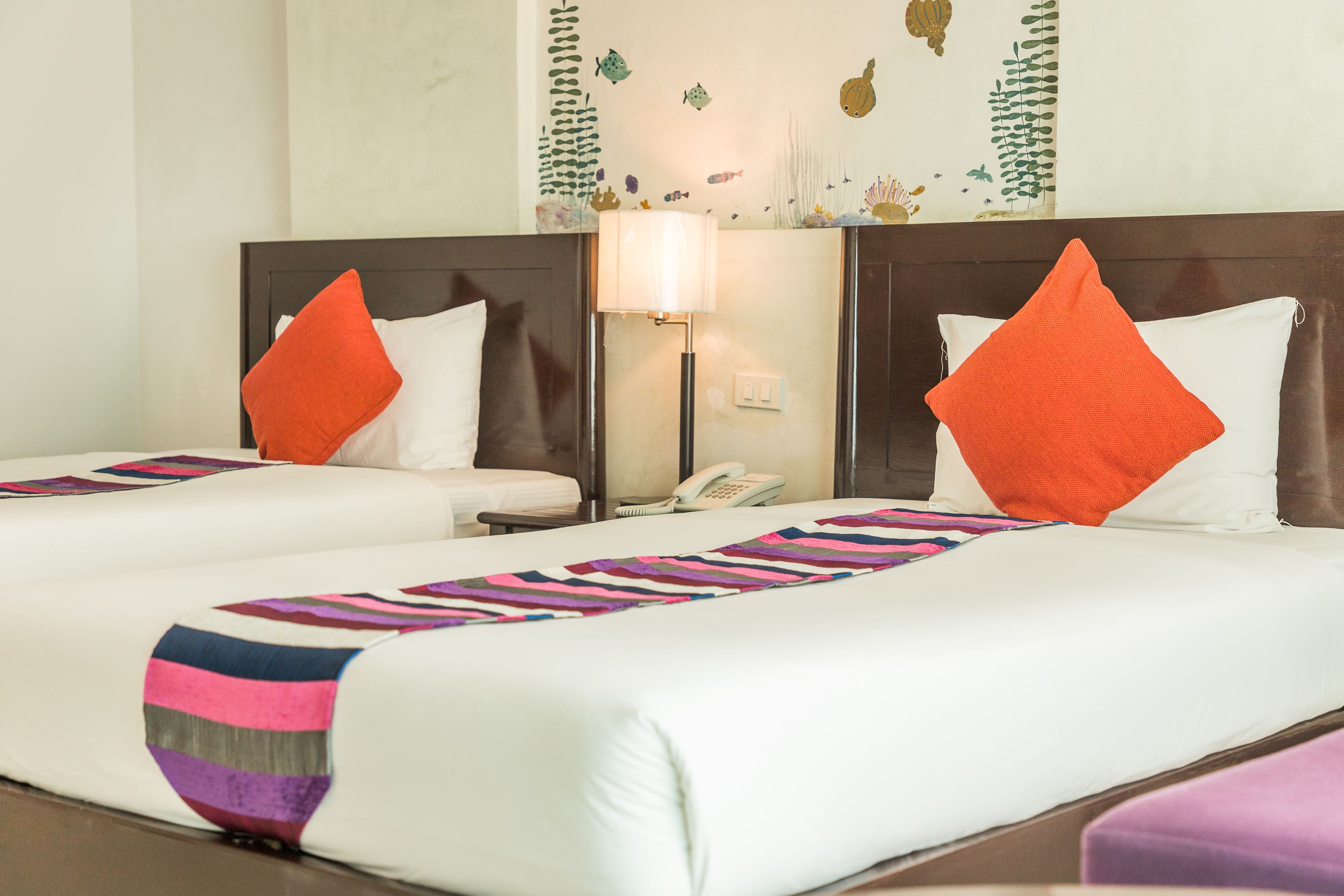 A hotel room with two beds, each with white linens and orange pillows, a nightstand with a lamp, and a wall mural of underwater scenes.