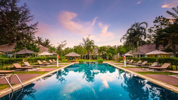 Une piscine entourée de chaises longues et de parasols, elle-même bordée de verdure et de bâtiments aux toits de chaume.