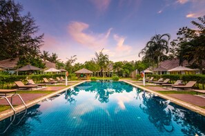 Ein Swimmingpool mit Liegestühlen und Sonnenschirmen, umgeben von Grünflächen und Gebäuden mit Strohdächern.