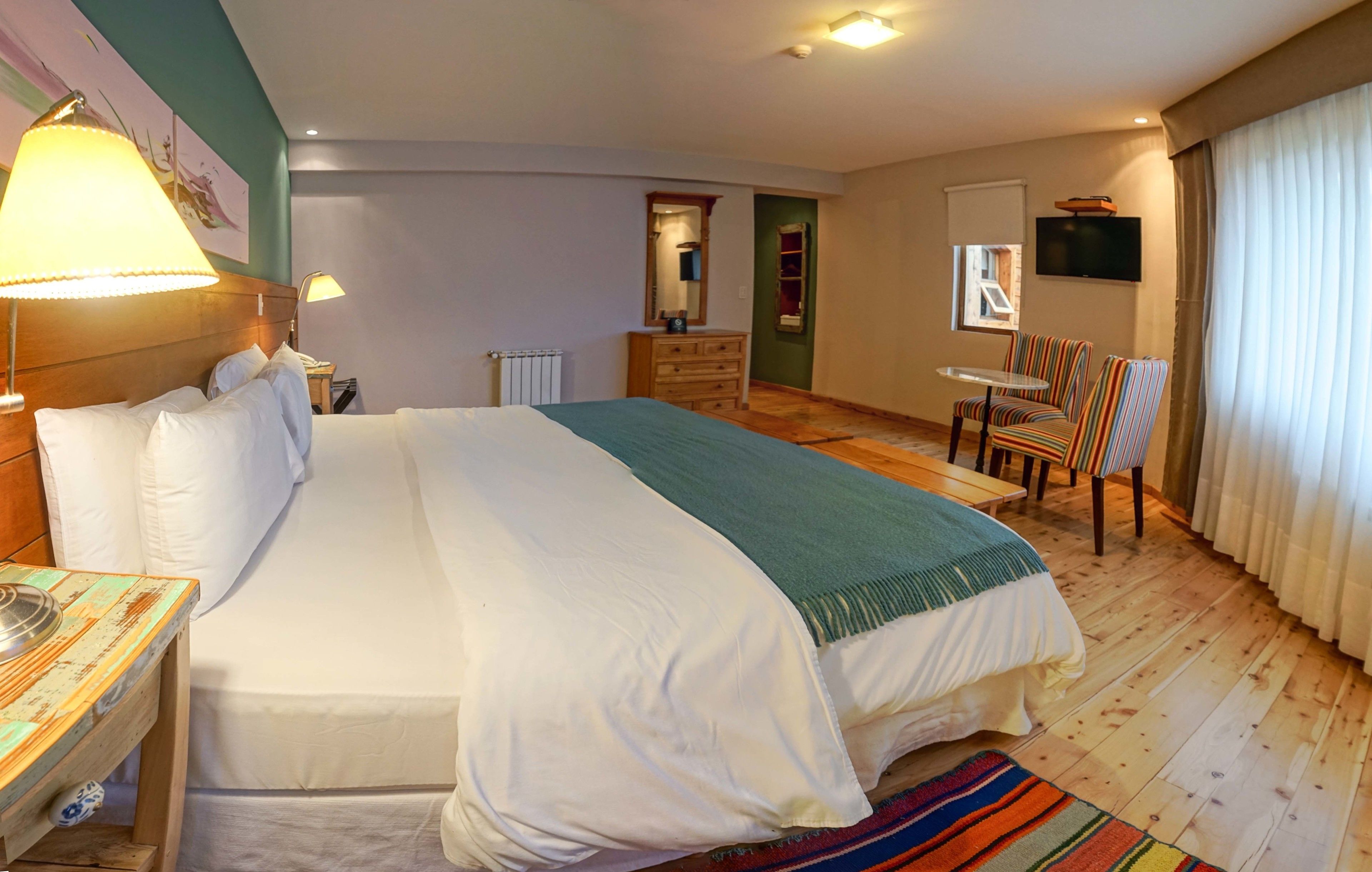 Quarto de hotel com uma cama grande, mesa de cabeceira de madeira, escrivaninha com luminária, uma cadeira, televisão e janela com cortinas.