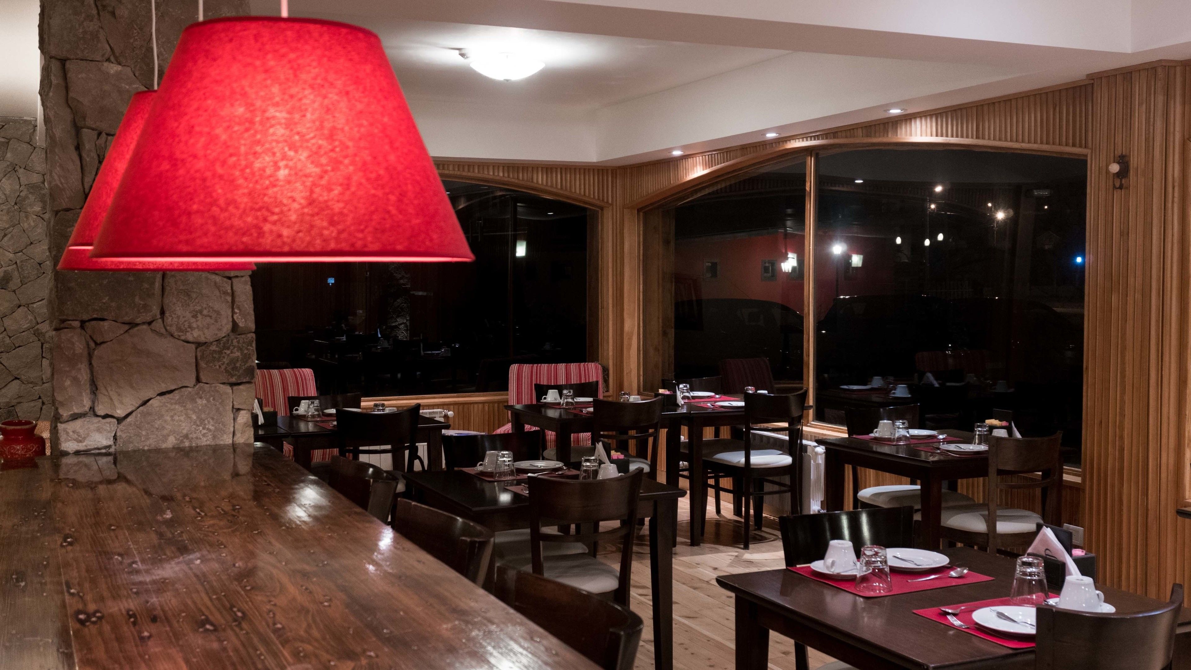 Un restaurante con mesas y sillas de madera, una lámpara colgante roja, paredes de piedra y ventanas amplias.