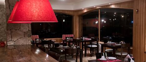 Un restaurante con mesas y sillas de madera, una lámpara colgante roja, paredes de piedra y ventanas amplias.