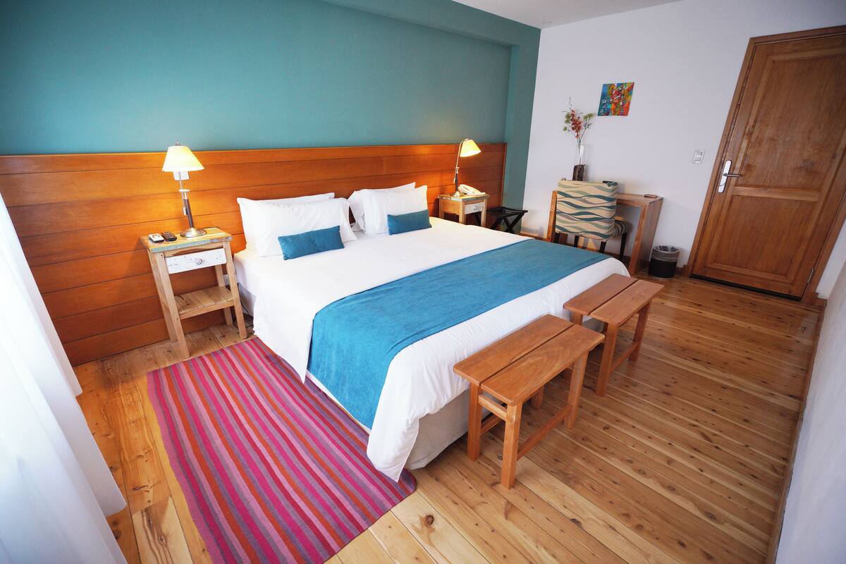 A bedroom with a wooden headboard, a bed with white and blue bedding, a bedside table with a lamp, a wooden bench, and a striped rug.