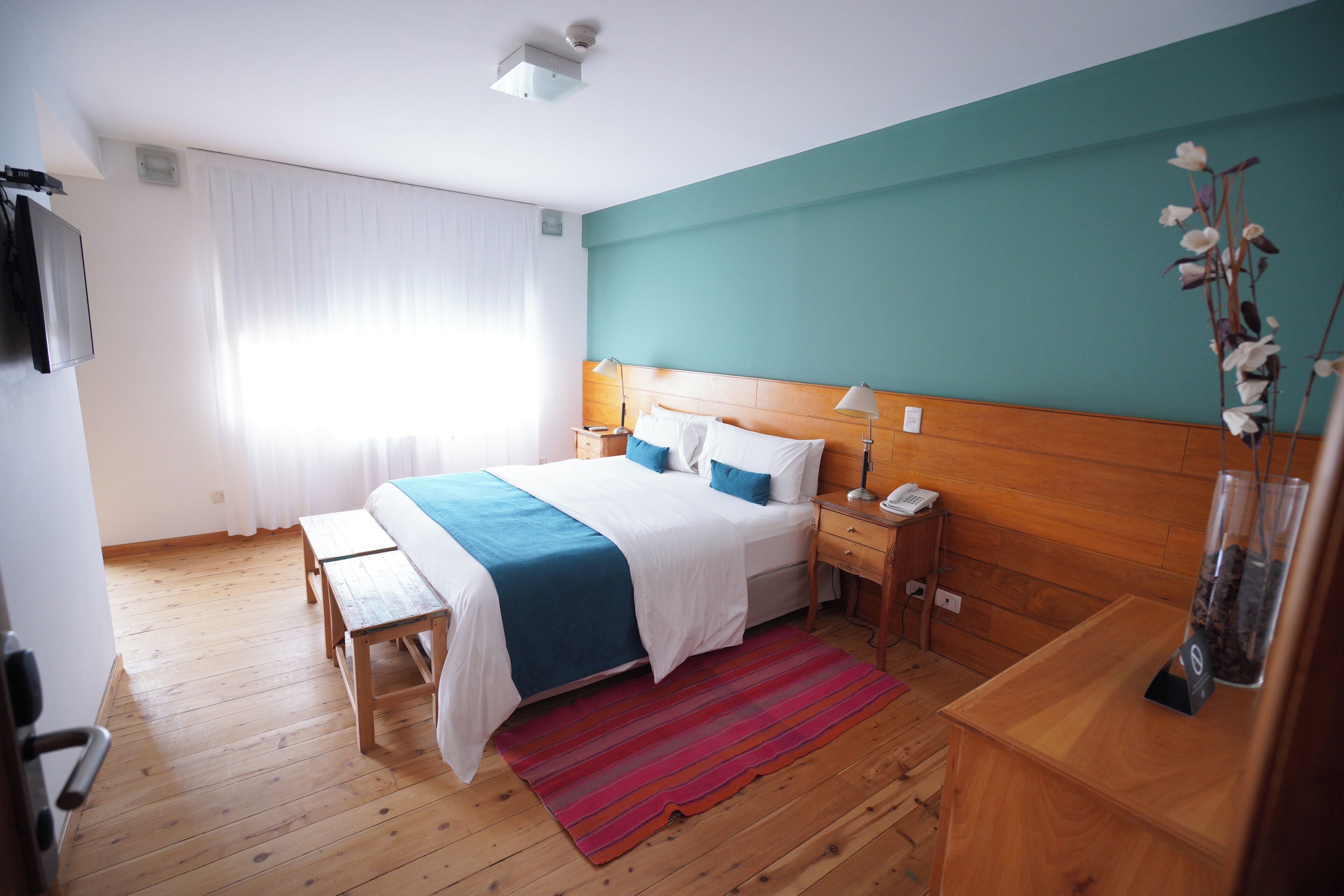 A hotel room with a wooden floor, a bed with a blue and white bedspread, a wooden nightstand, a desk, and a window with white curtains.