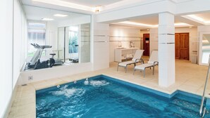 Uma área de piscina interior moderna, com estrutura de vidro, equipamentos de ginástica e espreguiçadeiras.
