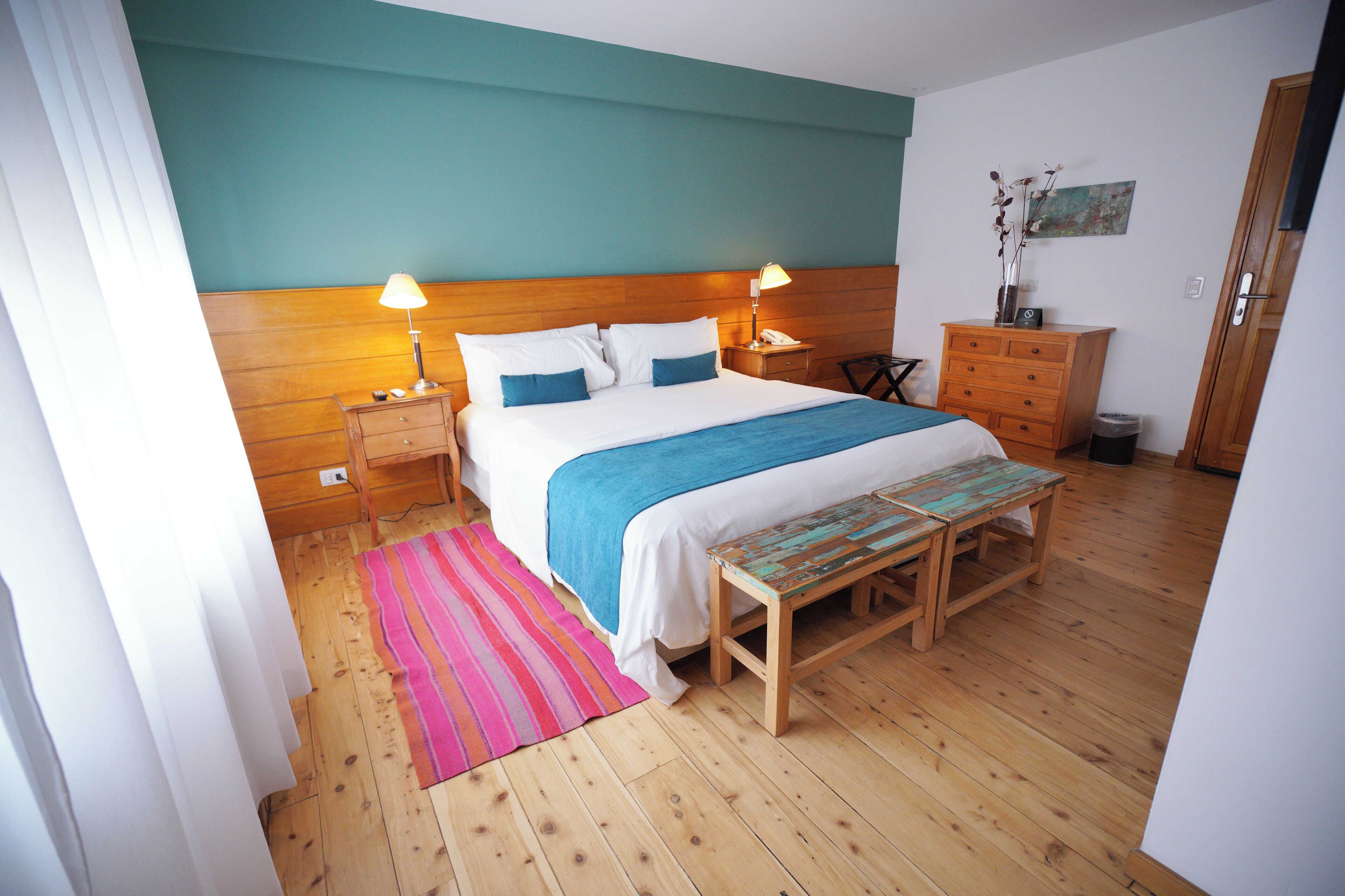 A bedroom with a wooden headboard, a bed with white and blue bedding, two bedside lamps, a wooden bench, a dresser, and a trash can.