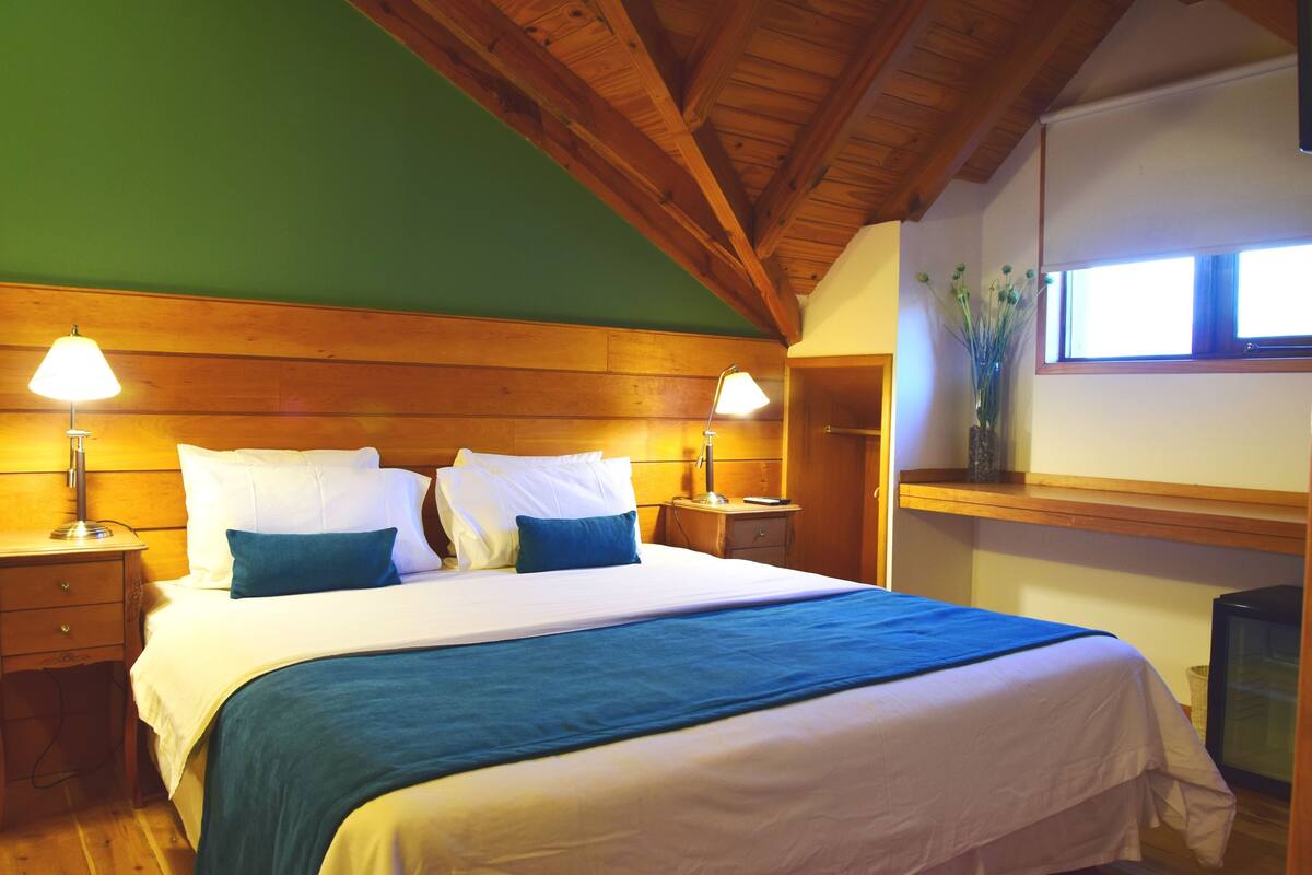A bedroom with a wooden ceiling, a bed with white and blue bedding, two bedside lamps, a nightstand, and a window with a view of greenery.