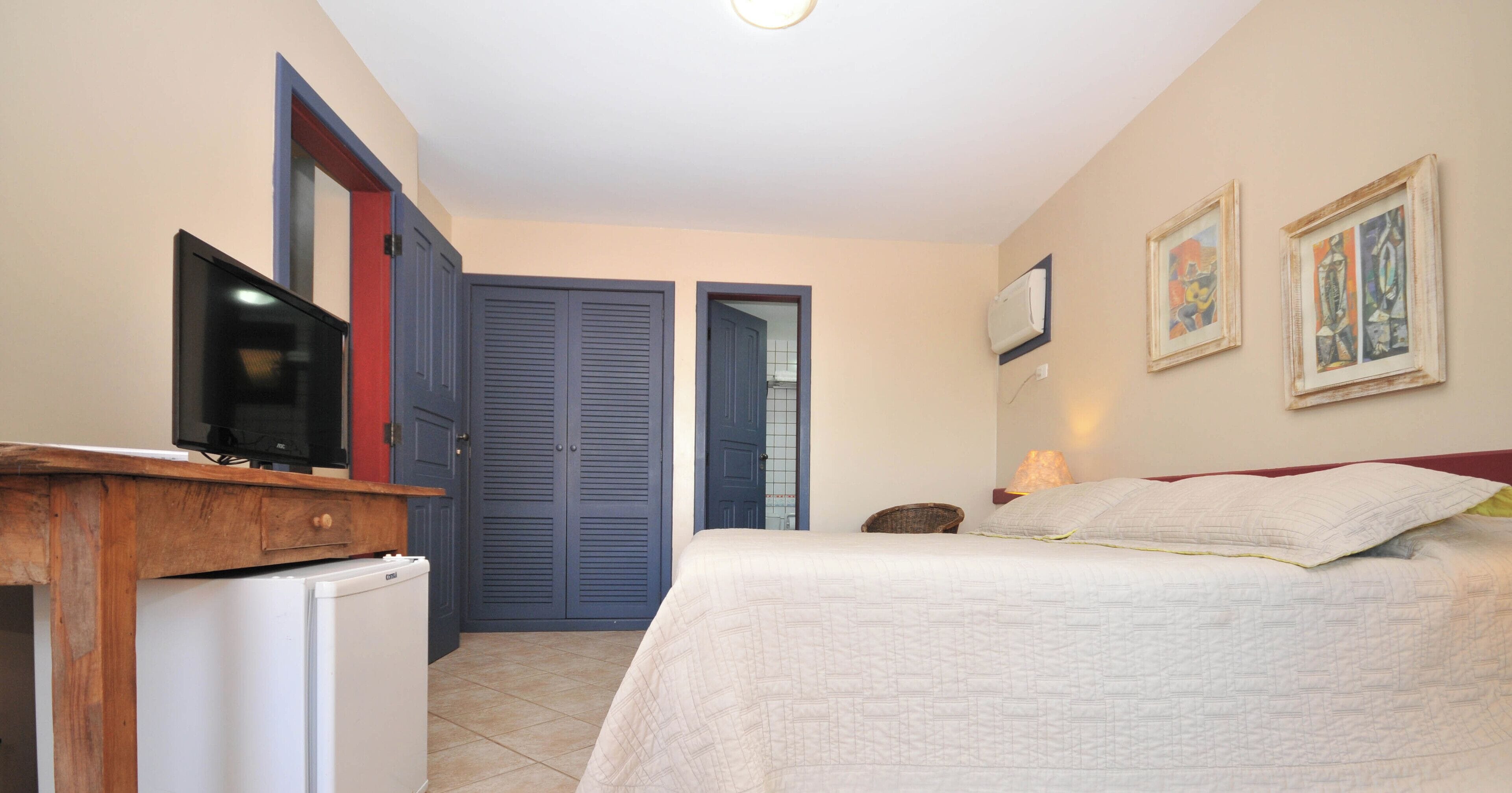 A bedroom with a bed, a television on a wooden desk, a wardrobe, and two framed pictures on the wall.