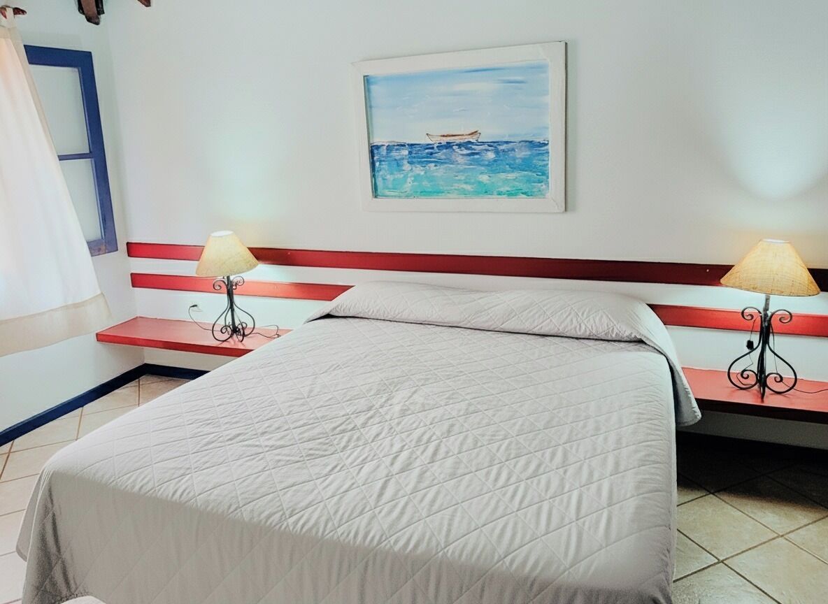 A hotel room with a bed, two bedside tables with lamps, a painting of a boat on water, and a red and white striped headboard.
