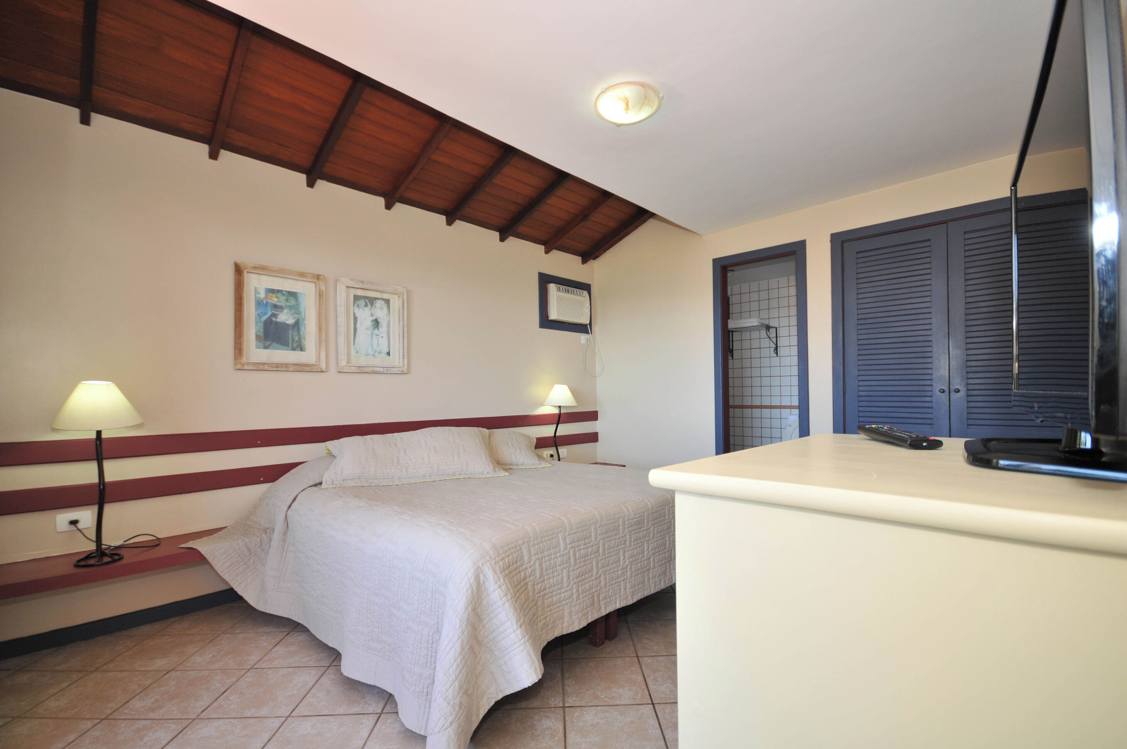 Une chambre d’hôtel avec un lit, deux lampes de chevet, un ventilateur de plafond, une télévision et une fenêtre avec des volets.
