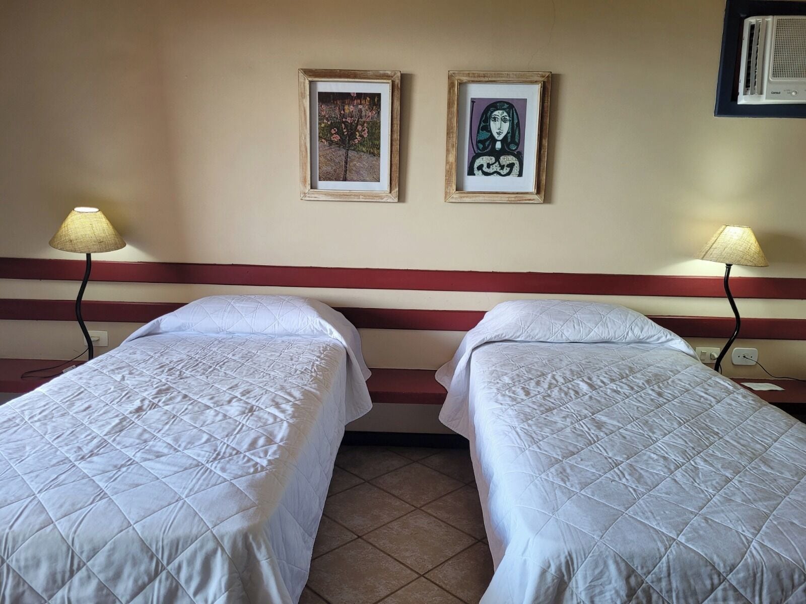 Habitació d'hotel amb dos llits, un capçal de llit vermell, dues fotografies emmarcades a la paret i vàries làmpades de nit.