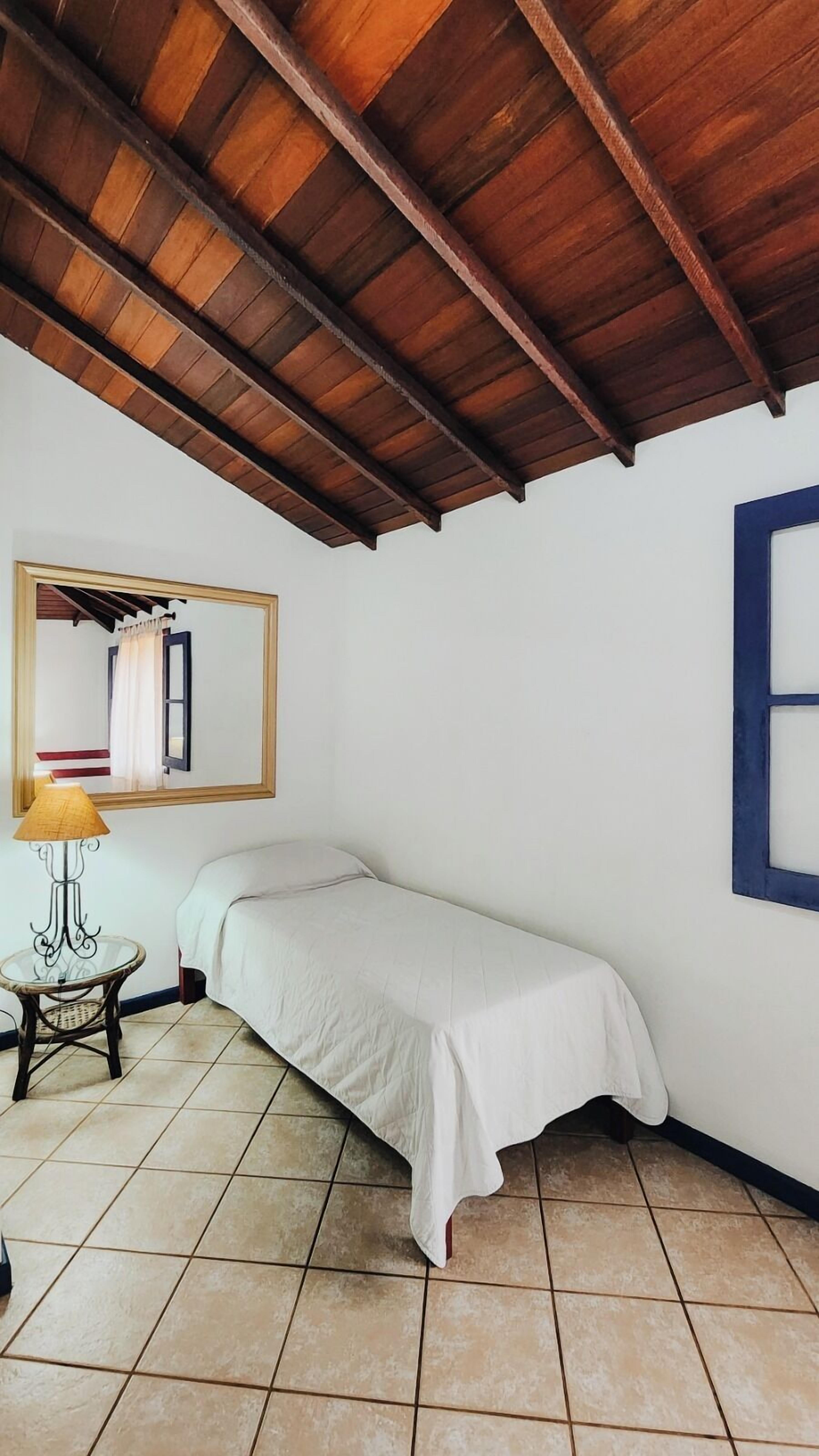 A small, single-bed room with a wooden ceiling, a mirror on the wall, a small table with a lamp, and a window with a blue frame.