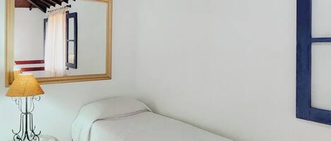 Una habitación pequeña con cama individual, techo de madera, un espejo en la pared, una mesita con lámpara y una ventana con marco azul.