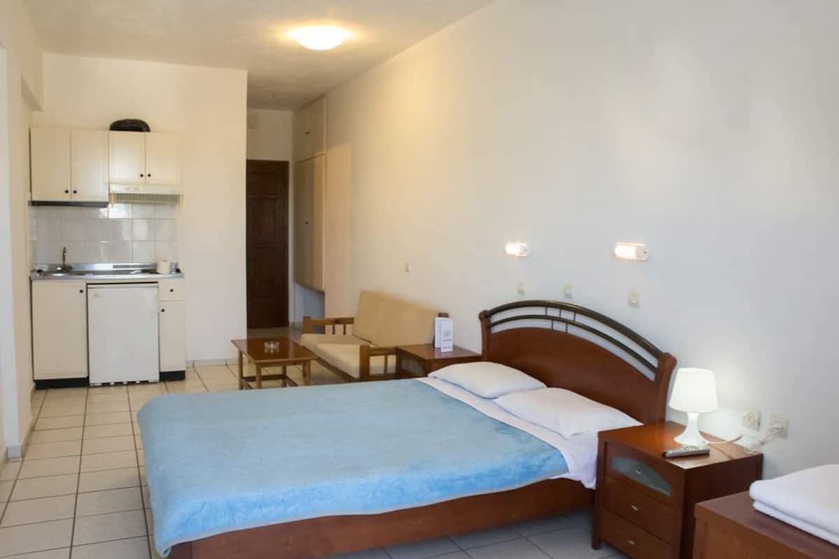 Een kleine hotelkamer met een eenpersoonsbed, een kitchenette, een stoel en een nachtkastje met een lamp.