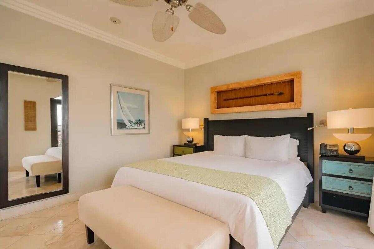 Une chambre d’hôtel avec un lit, des tables de chevet, un miroir, un ventilateur de plafond et un tableau encadré au mur.