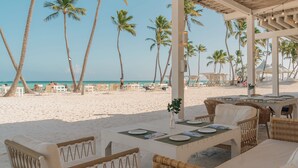 Un espace de restauration aménagé au bord de la plage, avec des meubles en osier, une table dressée pour deux personnes et une vue sur l’océan.