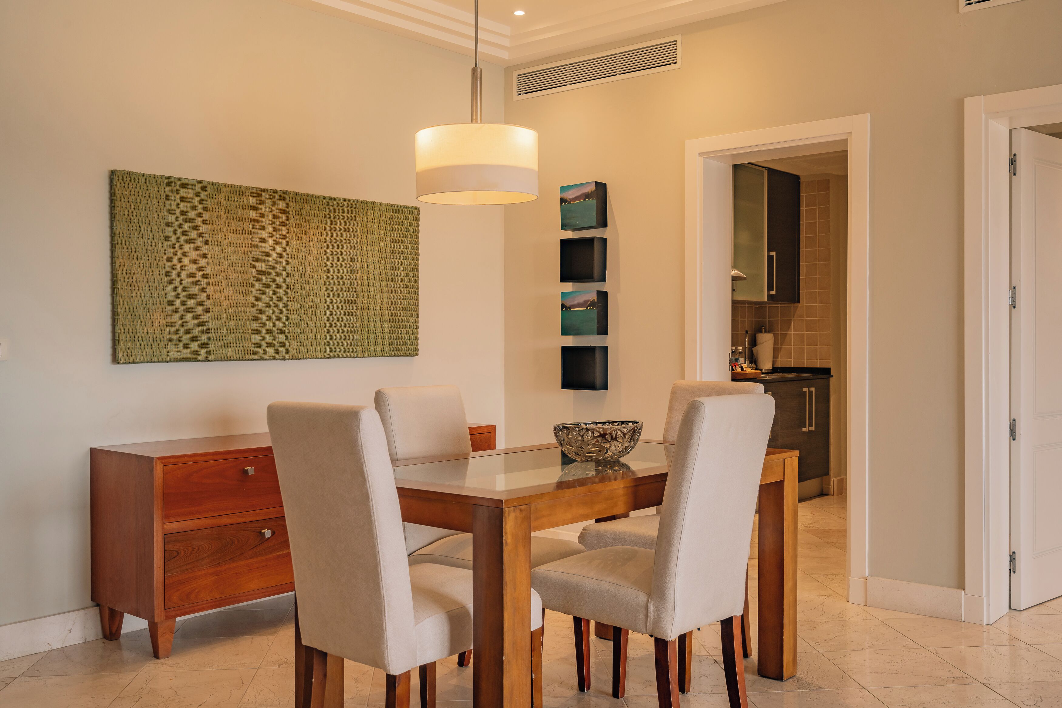 A dining area with a wooden table, four chairs, a glass-top table, a hanging light fixture, and a wall-mounted artwork.