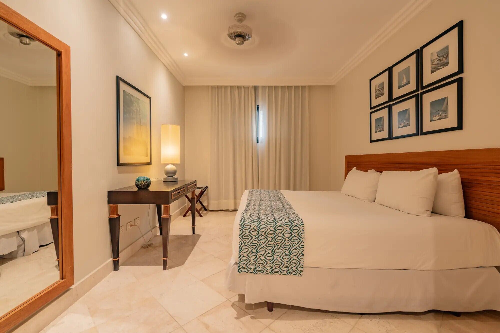 A hotel room with a bed, a desk, a lamp, a mirror, and framed pictures on the wall.