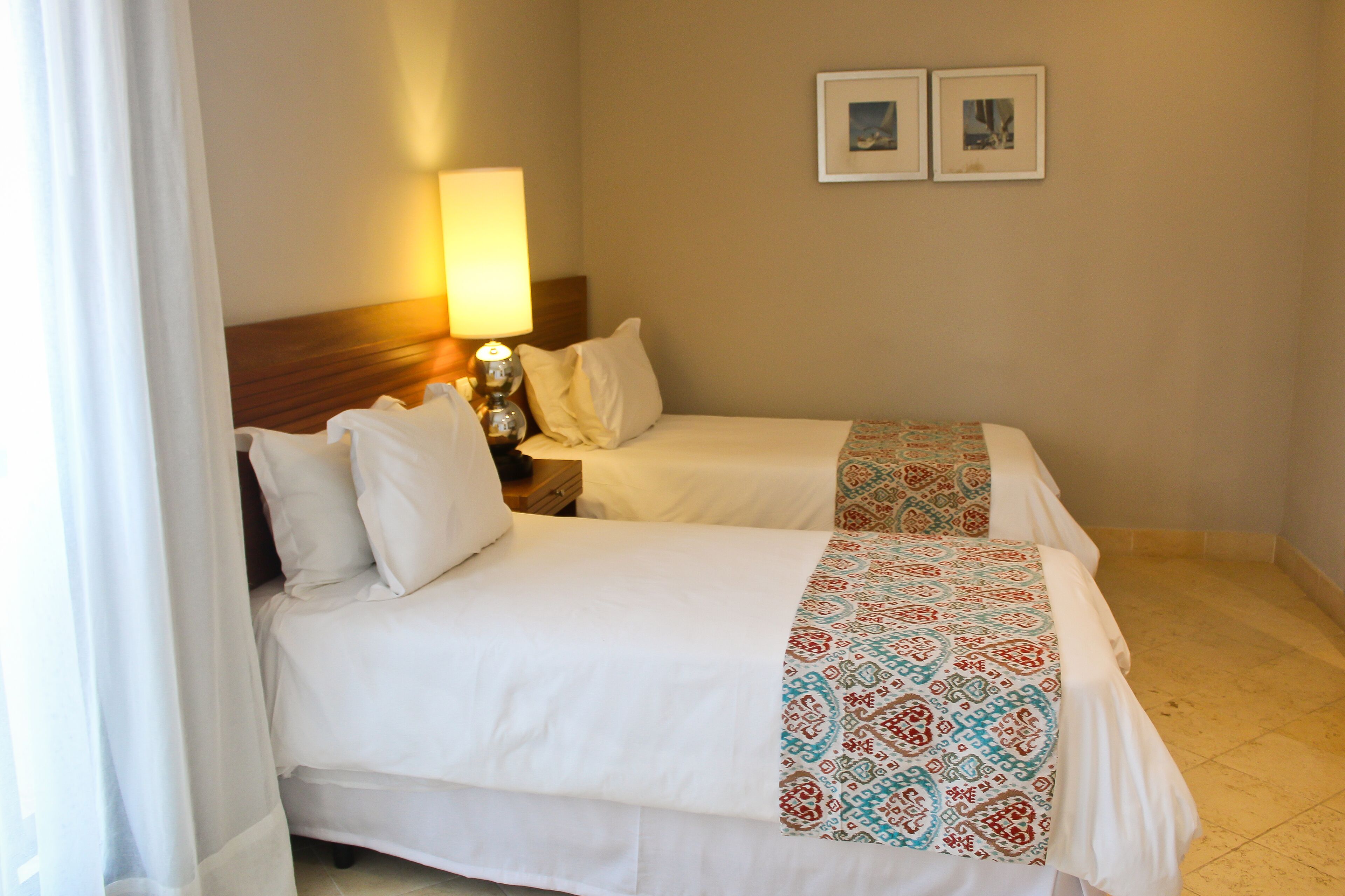 Una habitación de hotel con dos camas, una mesita de noche con lámpara y dos cuadros en la pared.