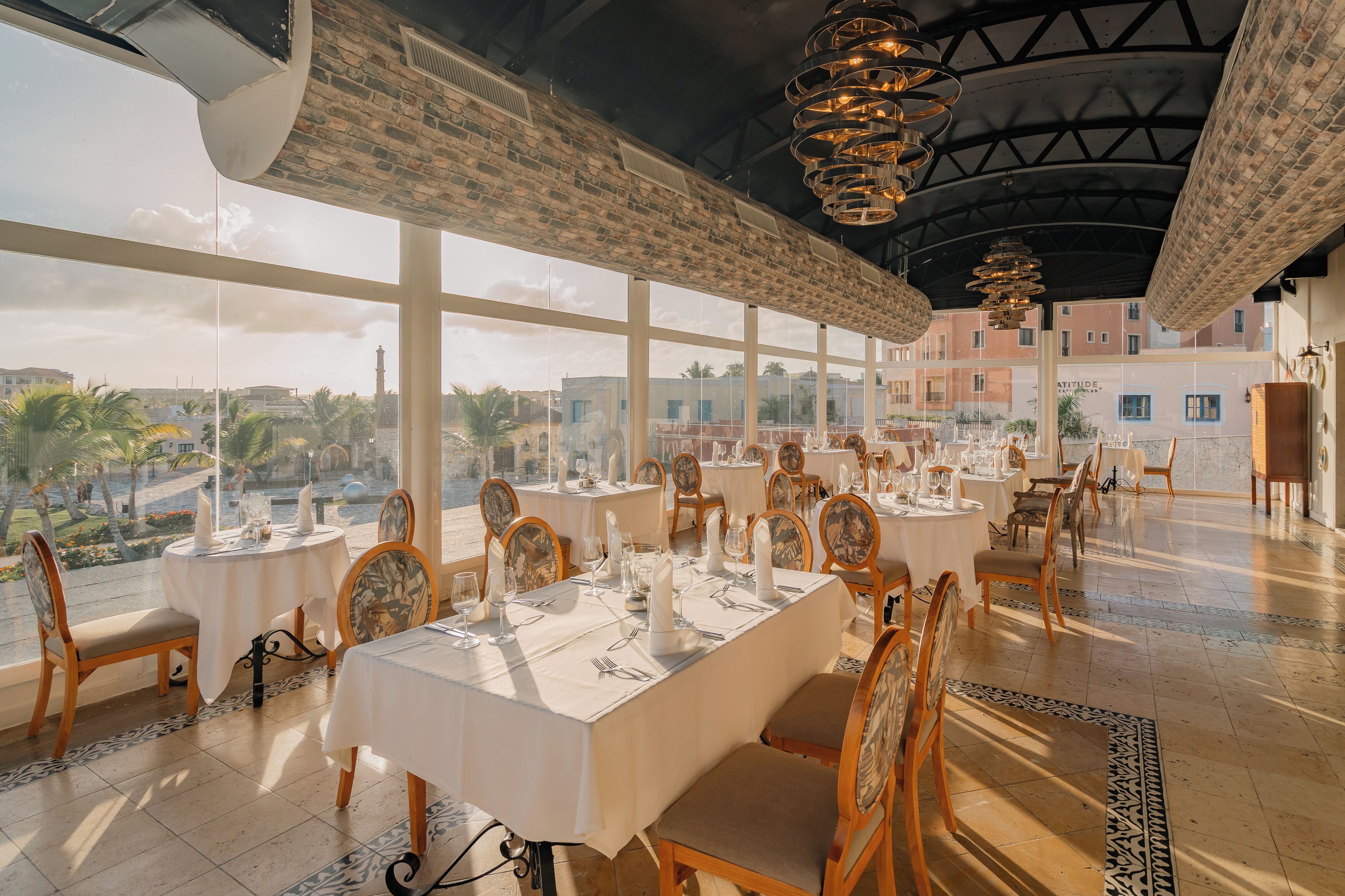 A restaurant with multiple tables set for dining, large windows offering a view of the city, and a high ceiling with decorative lighting.