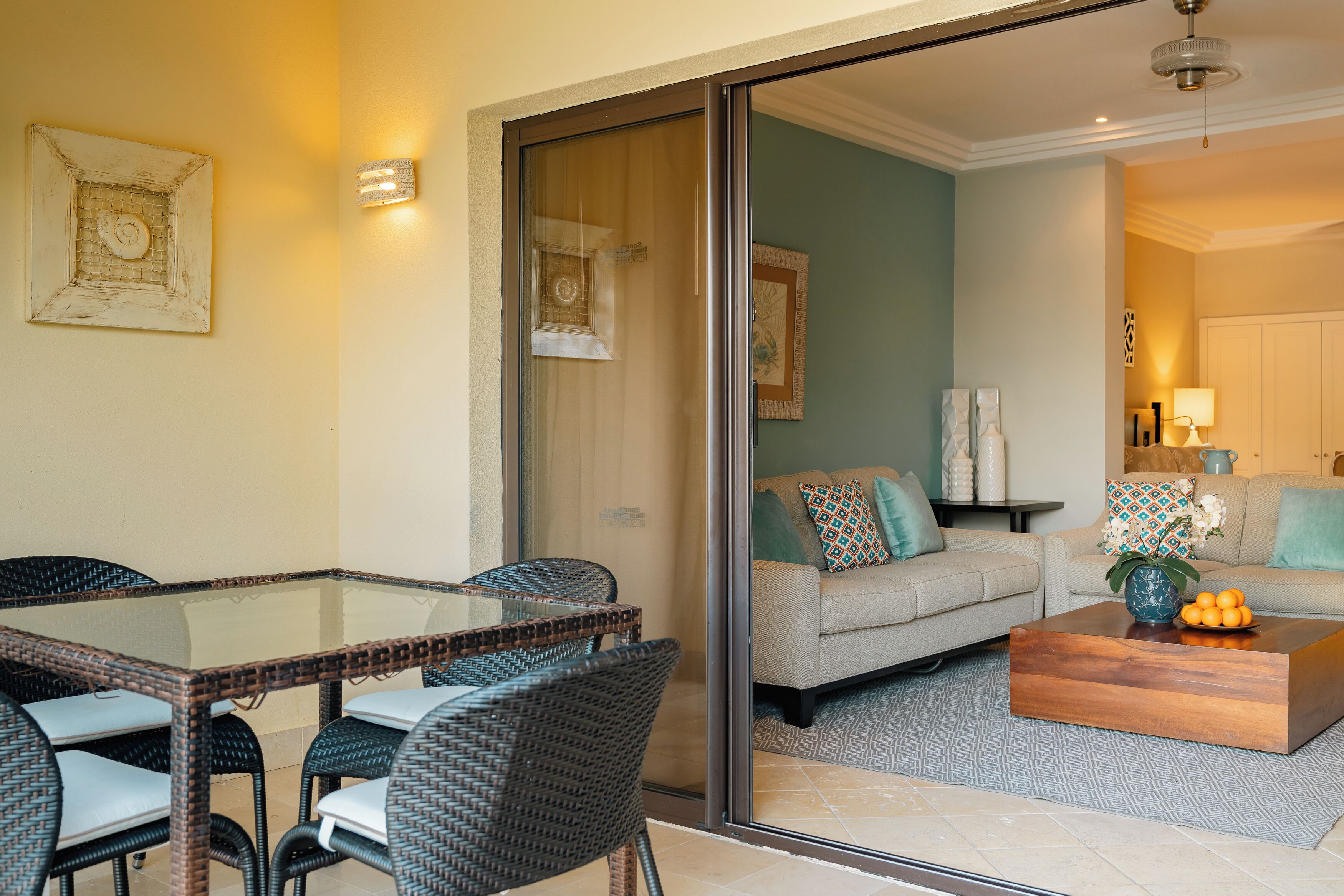 A living room with a glass partition, a sofa, a coffee table, and a dining area with wicker chairs.
