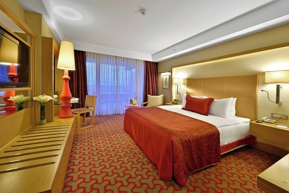 Ein Hotelzimmer mit Bett, Nachttisch, Lampe, Fernseher und einem Schreibtisch mit Blumen.