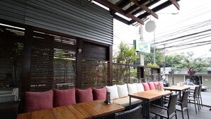 An outdoor seating area with wooden tables and wicker chairs, a covered section with wooden slats, and a street view with cars and buildings in the background.