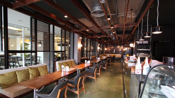 A modern restaurant interior with long wooden tables, cushioned seating, and large windows.