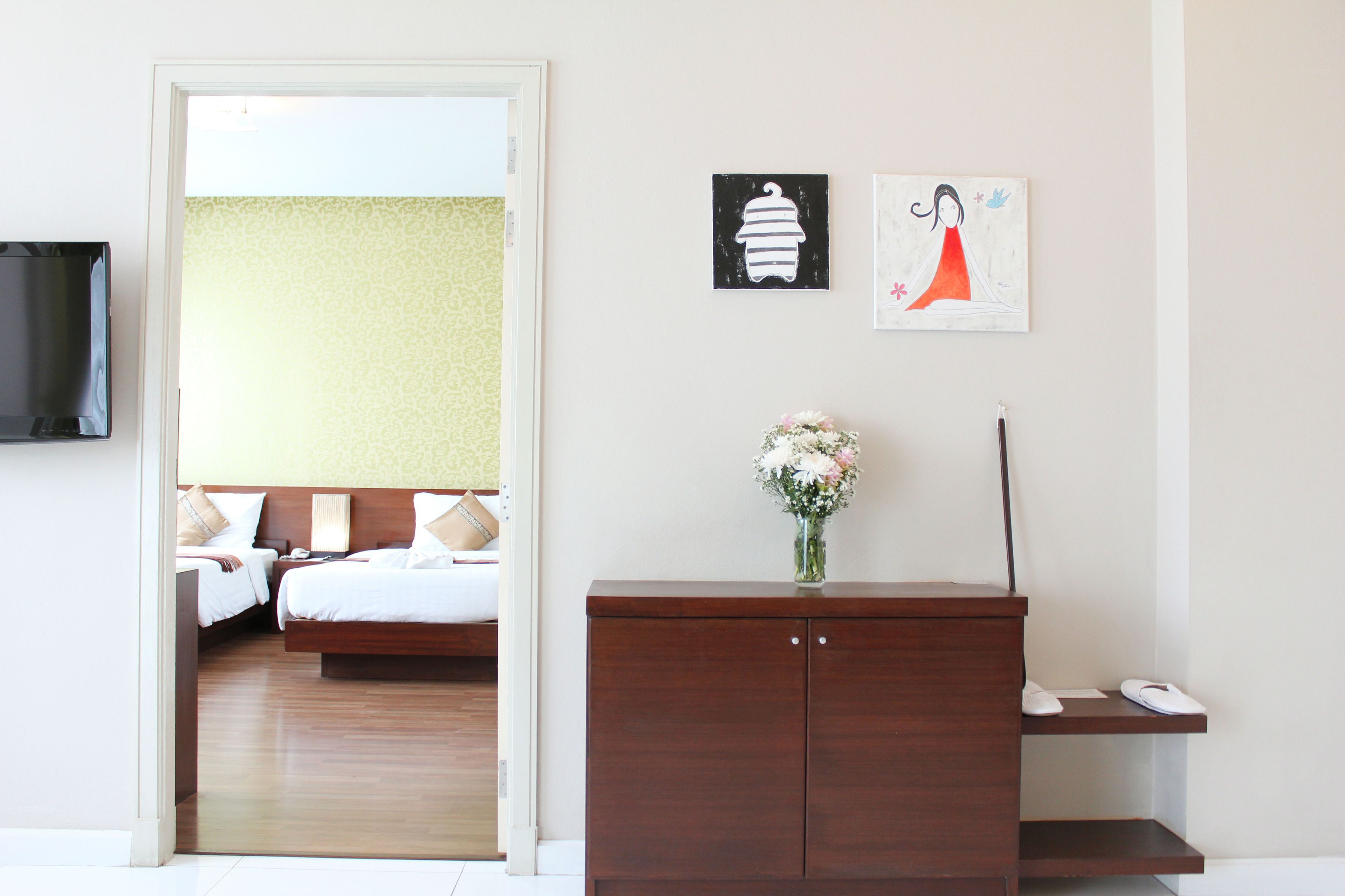 A hotel room with a wooden bed, a small cabinet, and a vase with flowers. There is a television on the wall and artwork above the cabinet.