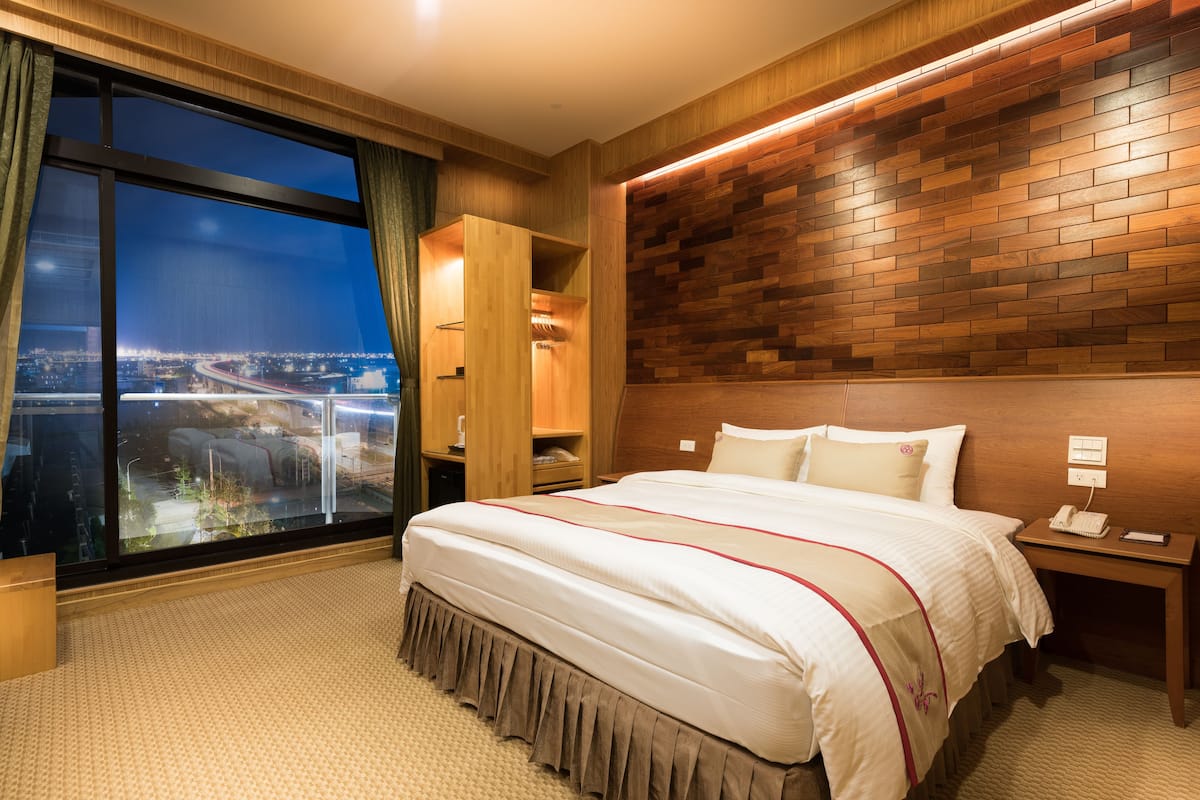 A hotel room with a large window, a bed with a wooden headboard, a nightstand, and a wardrobe.