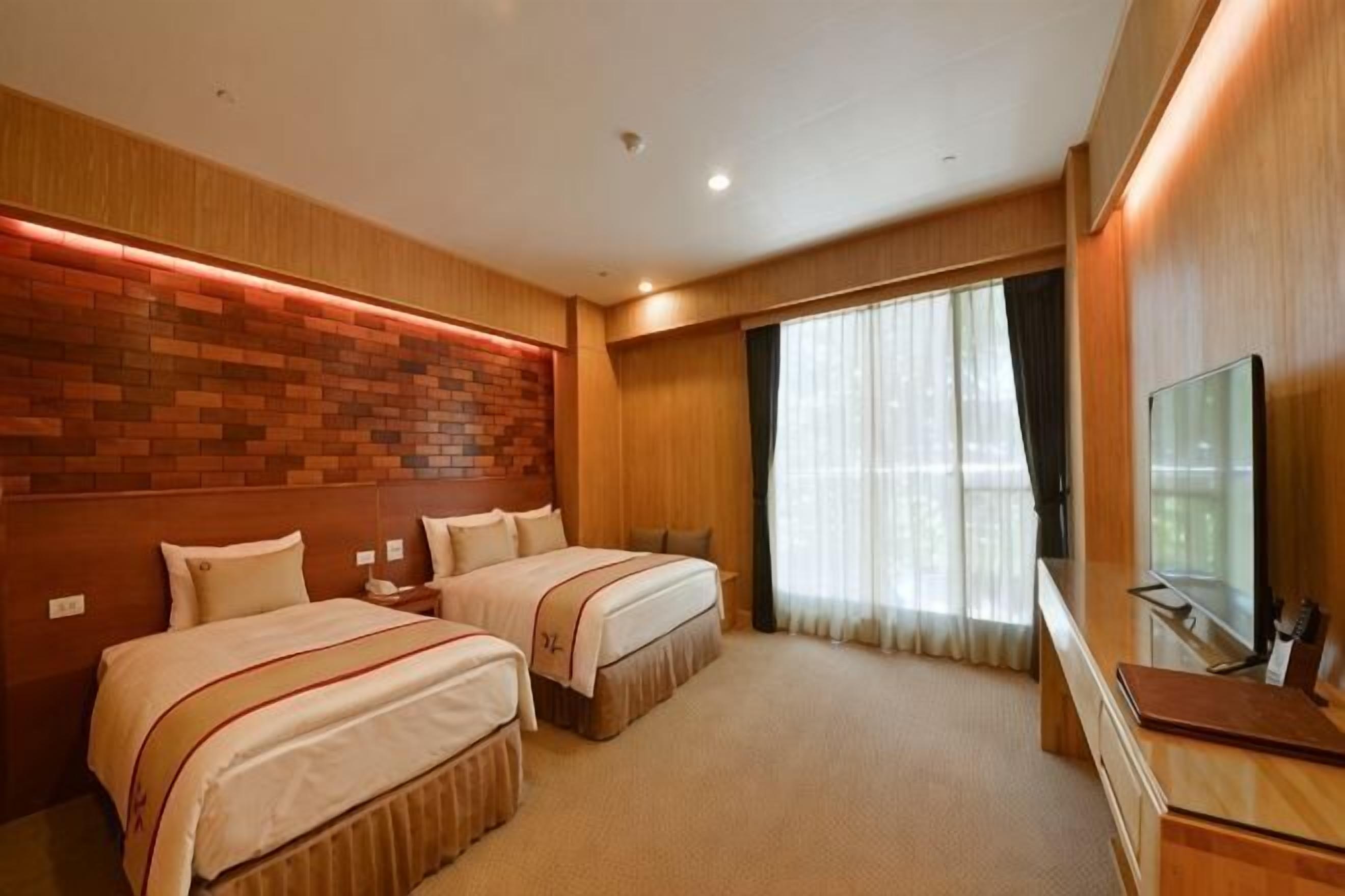 A hotel room with two beds, a TV, a desk, and a large window with curtains.