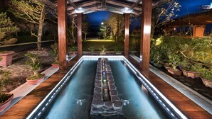 Una piscina rectangular con borde de piedra, rodeada de plantas en macetas y una estructura de pérgola de madera.