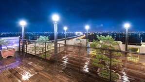 Una terraza de madera mojada con barandillas y plantas en macetas, con vista a un paisaje urbano nocturno.