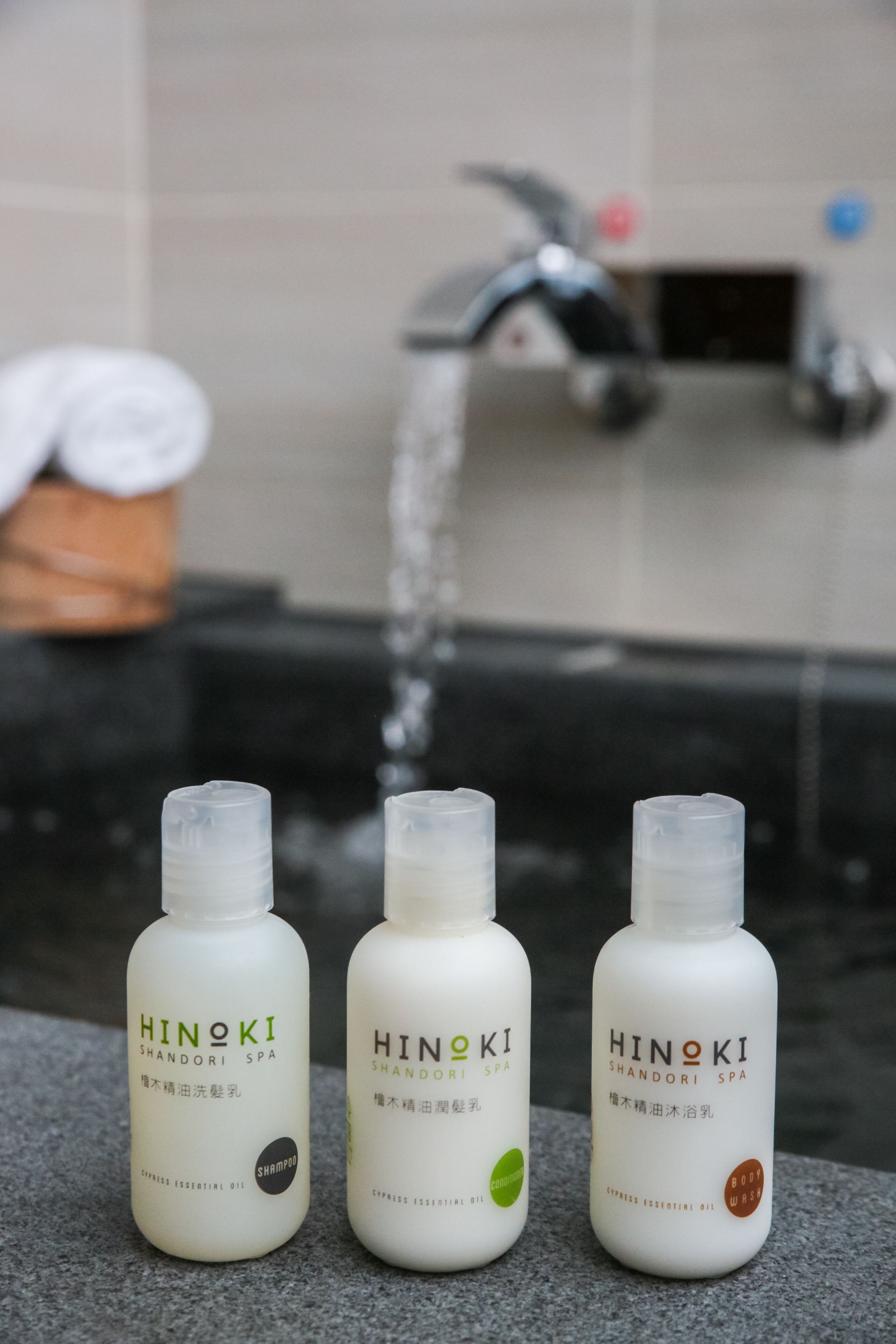 Tres botellas de Hinoki Shandori Spa sobre una superficie de piedra, con un grifo al fondo.