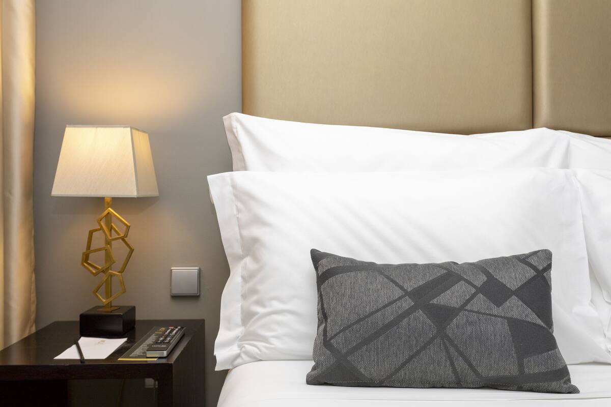 Un lit bien fait, avec du linge de lit blanc et un oreiller à motifs gris, une lampe de chevet au design géométrique et une petite table sur laquelle reposent une télécommande et un bloc-notes.