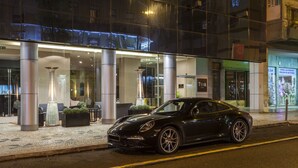 Un deportivo negro estacionado frente a un edificio moderno con fachada de vidrio, pilares grandes y una zona de estar al aire libre.