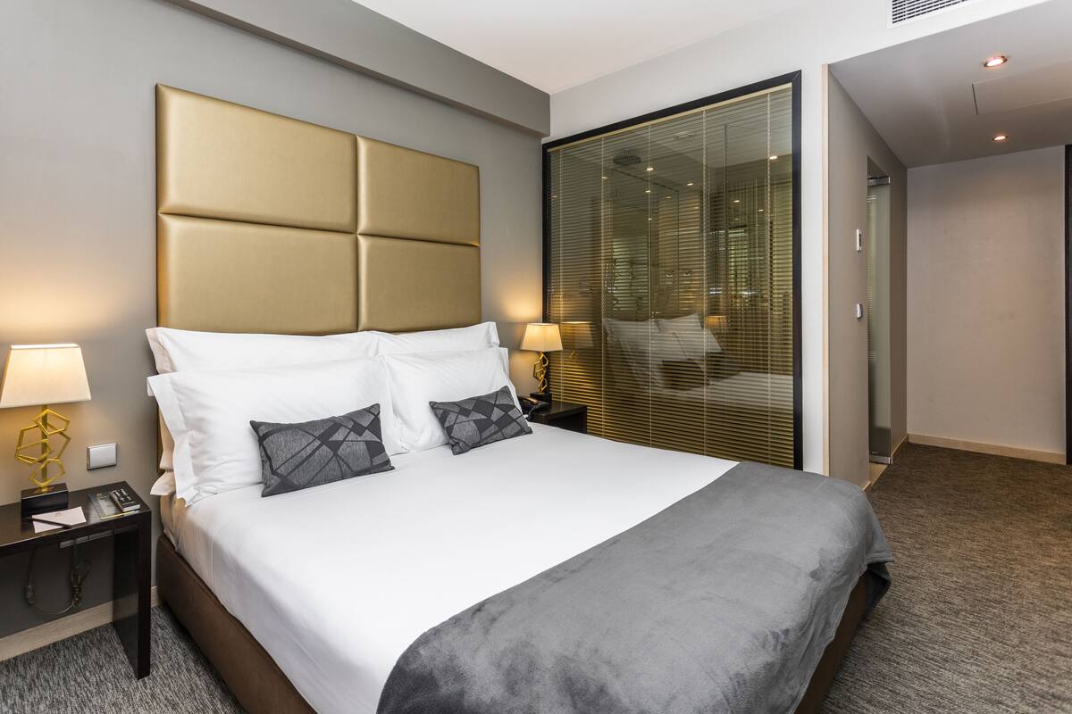 Une chambre d’hôtel moderne avec un grand lit, une table de chevet avec une lampe, une cloison en verre et un sol recouvert de moquette.