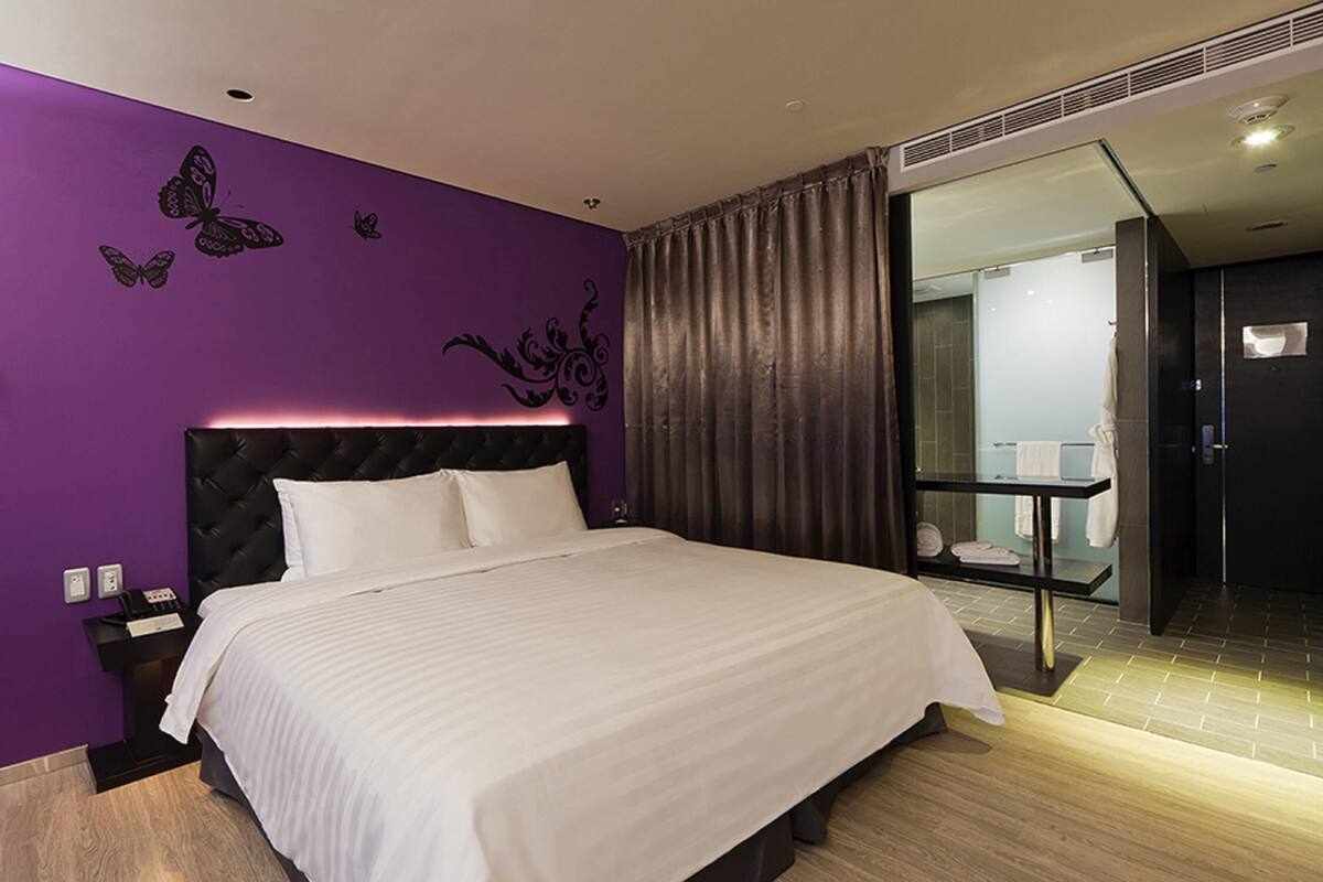 A modern hotel room with a large bed, purple walls, and a bathroom visible through an open door.