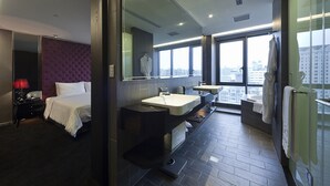Una habitación de hotel moderna con una cama grande, un tocador con espejo, dos lavabos y vistas a la ciudad a través de amplias ventanas.