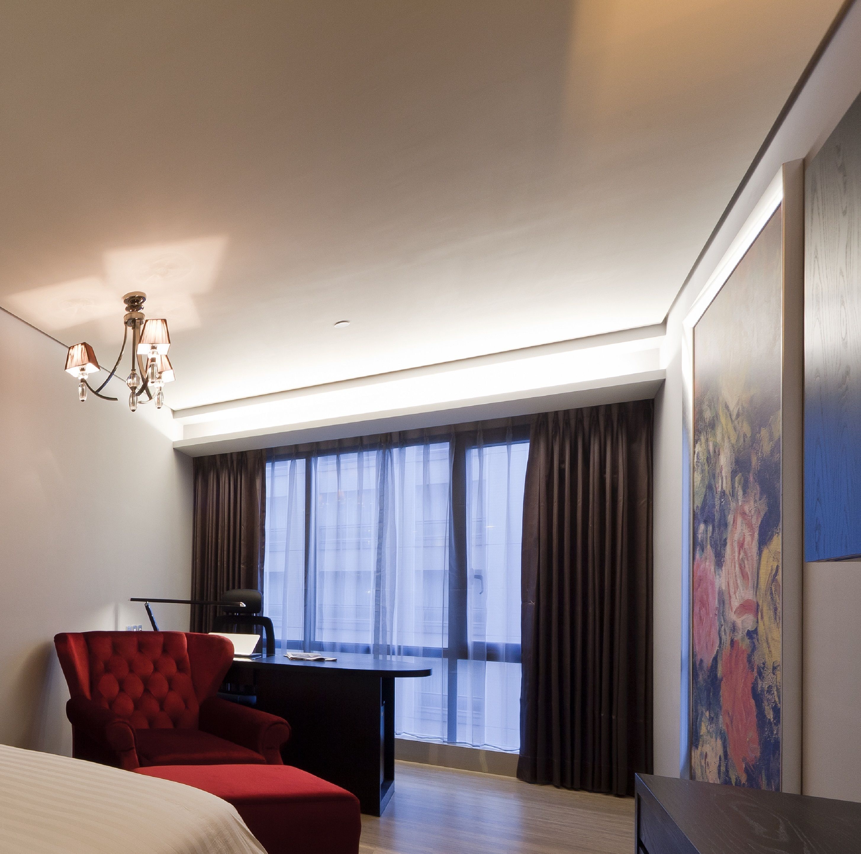 A modern hotel room with a large bed, a red armchair, a desk with a chair, a chandelier, and a large window with curtains.