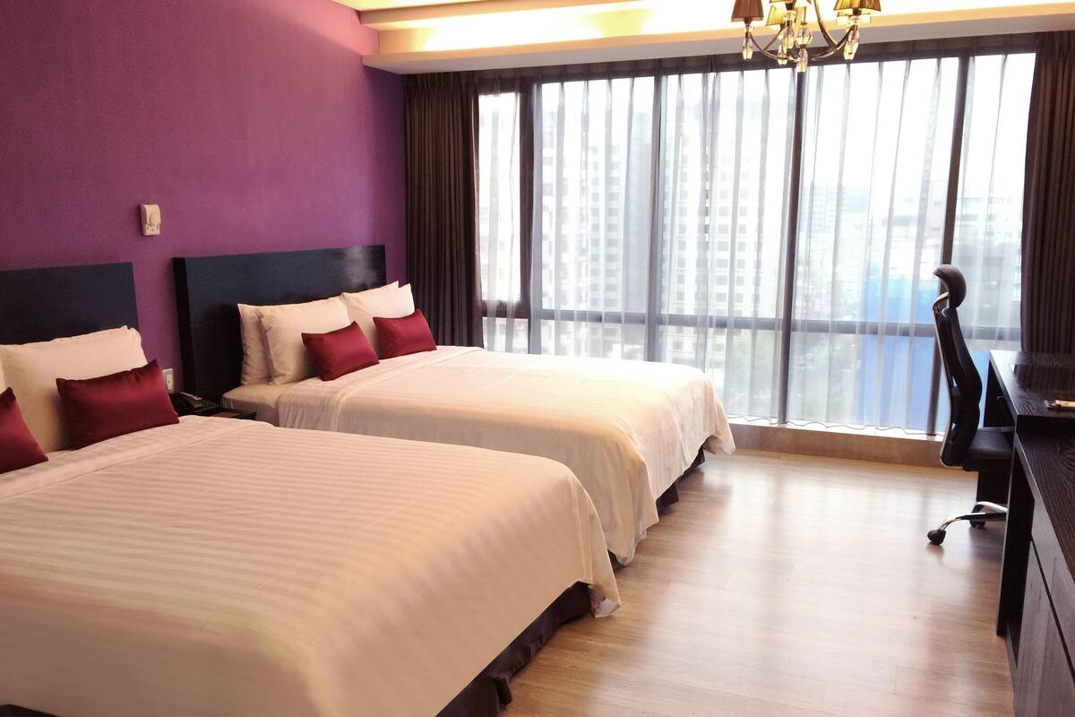 A hotel room with two beds, a desk, and a large window with a city view.