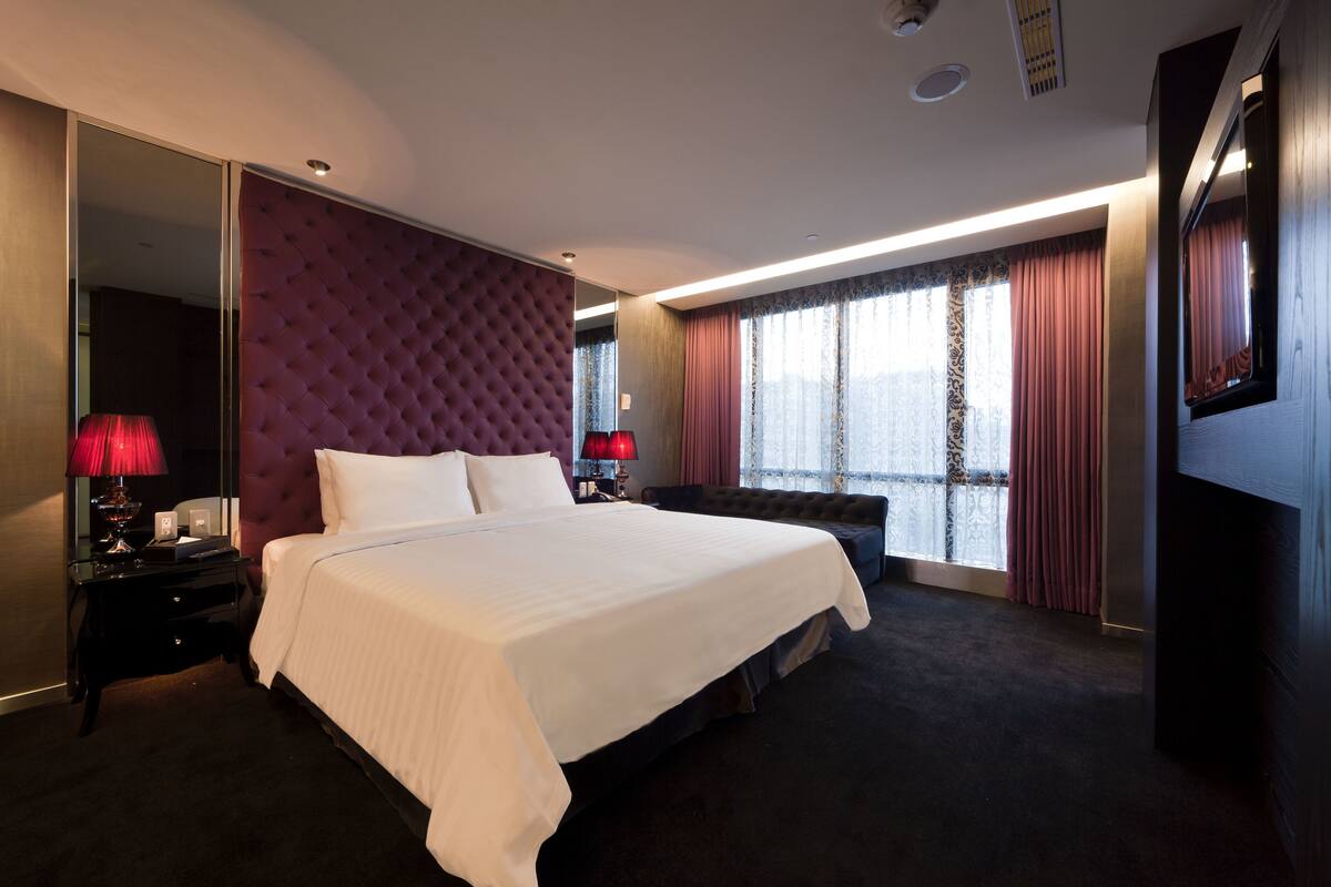 A large bed with white linens, a red upholstered headboard, and a dark carpeted floor. There is a large mirror on one wall and a flat-screen TV on another.