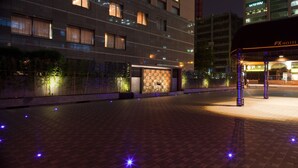 Entrada de hotel con diseño moderno, una zona de espera techada y un camino bien iluminado.