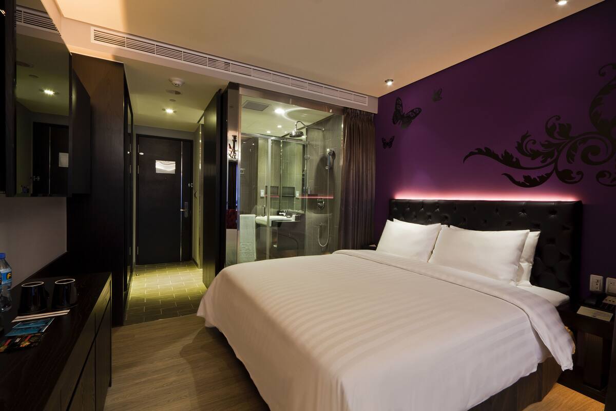 A modern hotel room with a large bed, a glass-enclosed shower, and a purple accent wall.