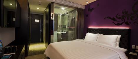 A modern hotel room with a large bed, a glass-enclosed shower, and a purple accent wall.